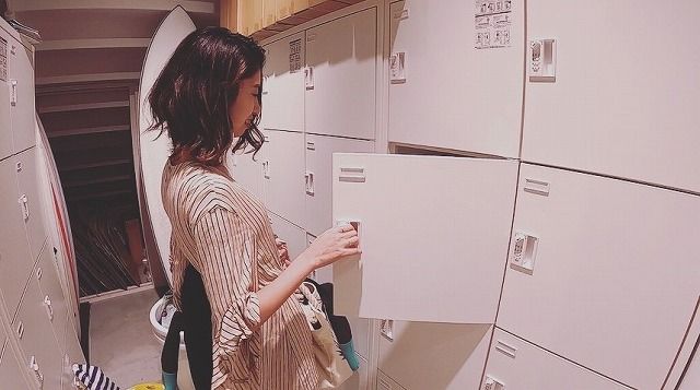 Woman with dark hair in a striped shirt opens a white storage locker in a row of lockers in a laundry room.