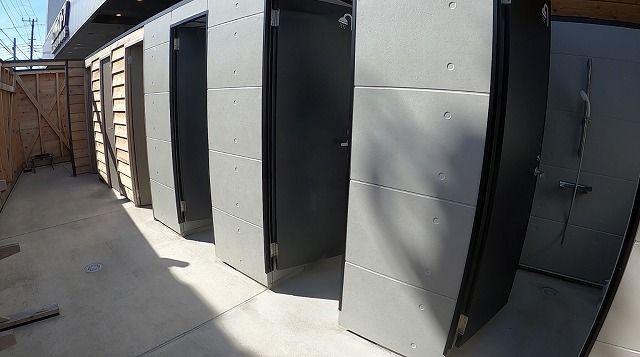 Row of grey metal storage unit doors along a concrete alley, with a wooden fence on the left and sunlight casting shadows on the ground.