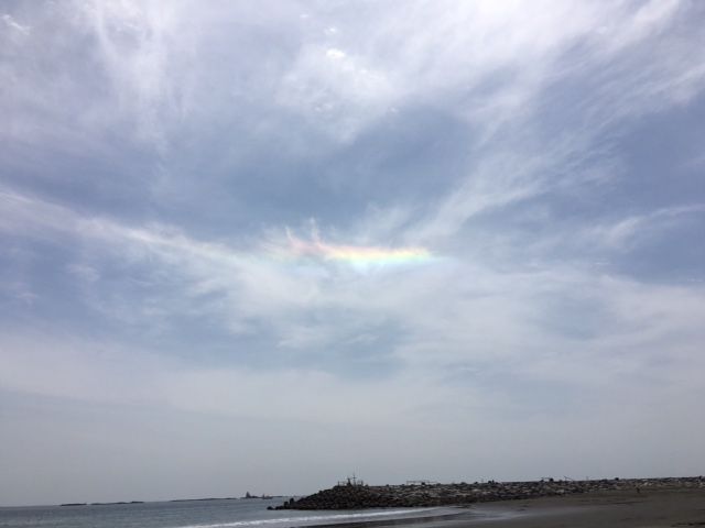 Sun halo rainbow in a pale blue sky over a calm sea with a rocky breakwater in the distance.