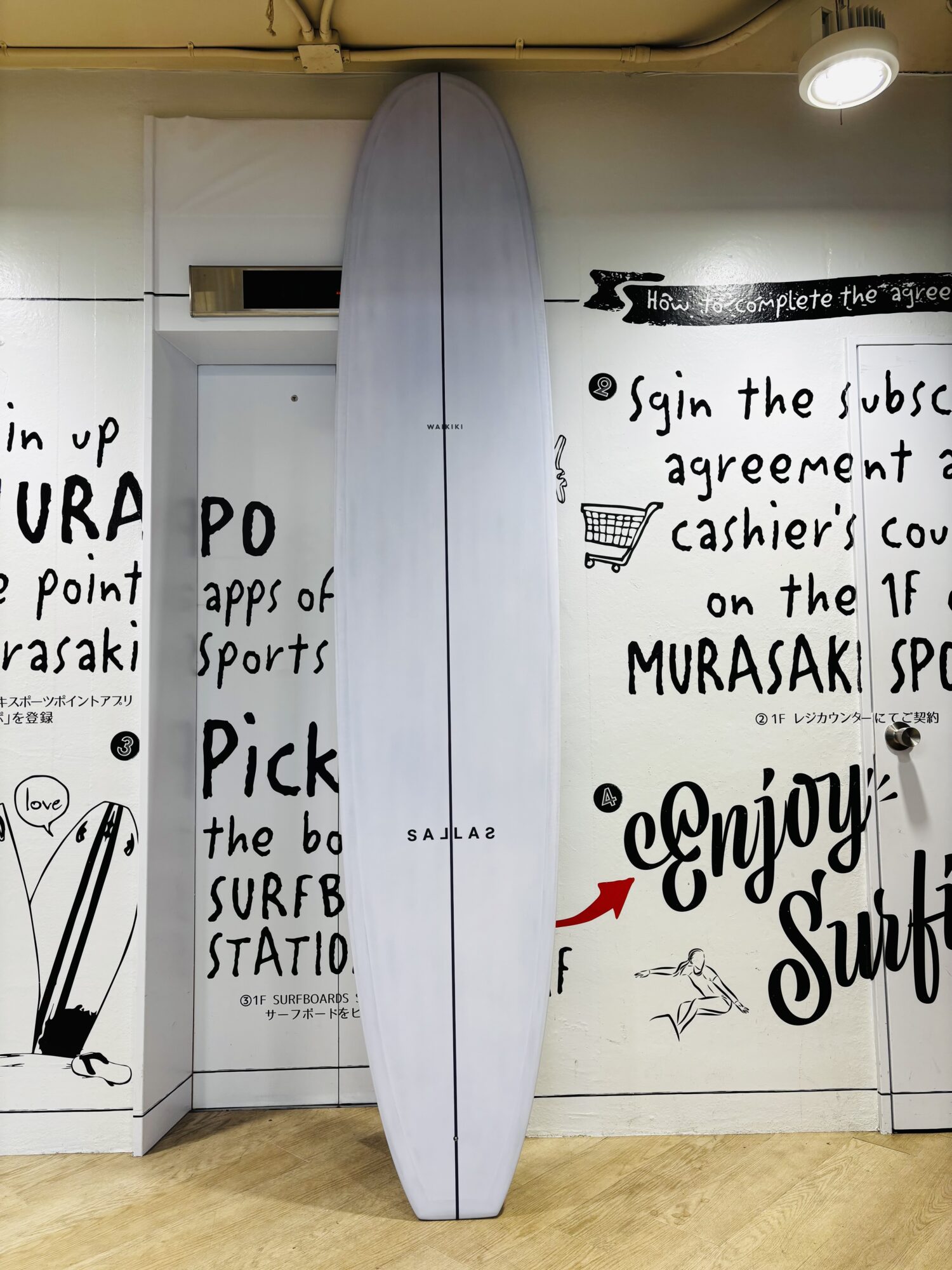 White surfboard standing upright between walls covered with bold black handwritten text and small drawings, on a wooden floor.