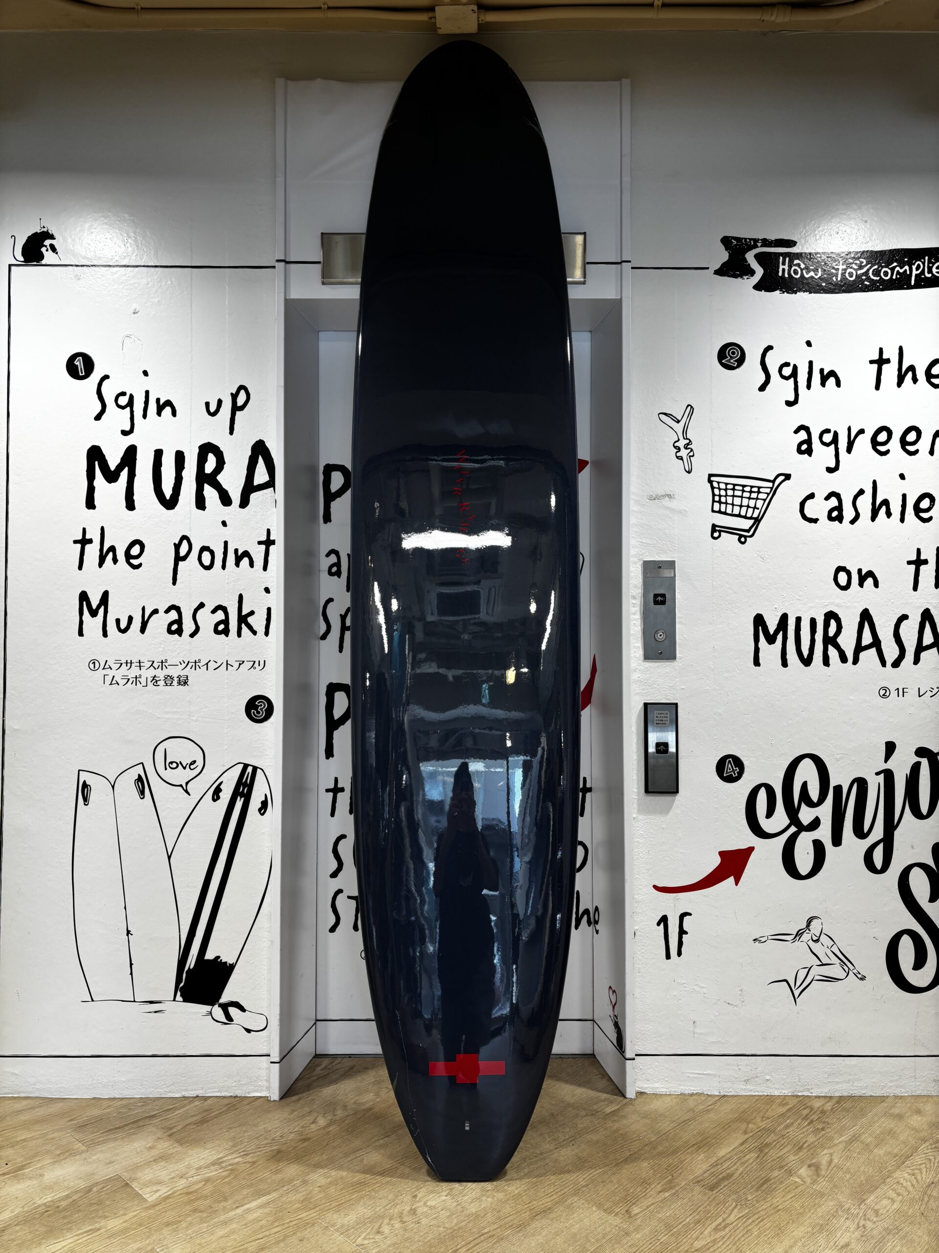 IMG_1128 - SHOP NEWS | ムラサキスポーツオンラインストア 通販 Tall glossy black surfboard standing upright in a white-walled display alcove with graffiti-style text and drawings on either side, and a wooden floor beneath.