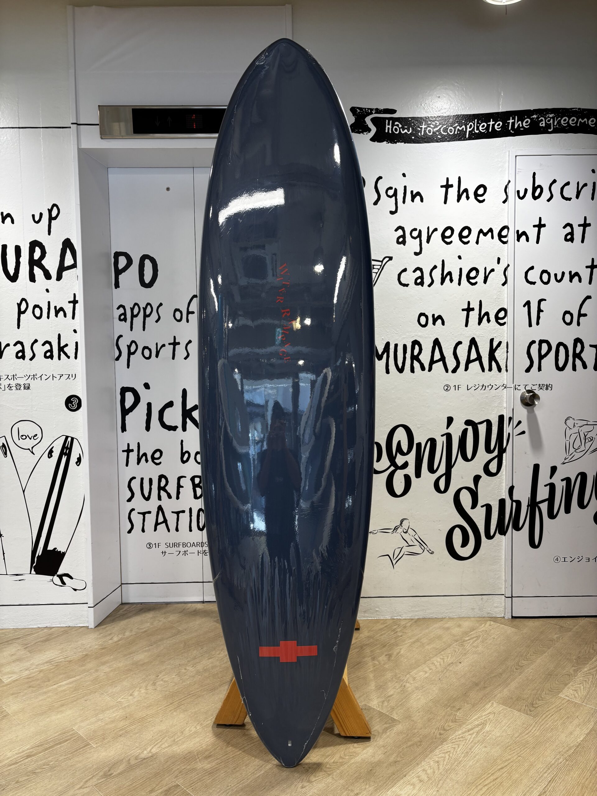 IMG_1132 - SHOP NEWS | ムラサキスポーツオンラインストア 通販 Blue surfboard standing upright on a wooden support indoors, with black handwritten wall text in the background and a red cross near the bottom.