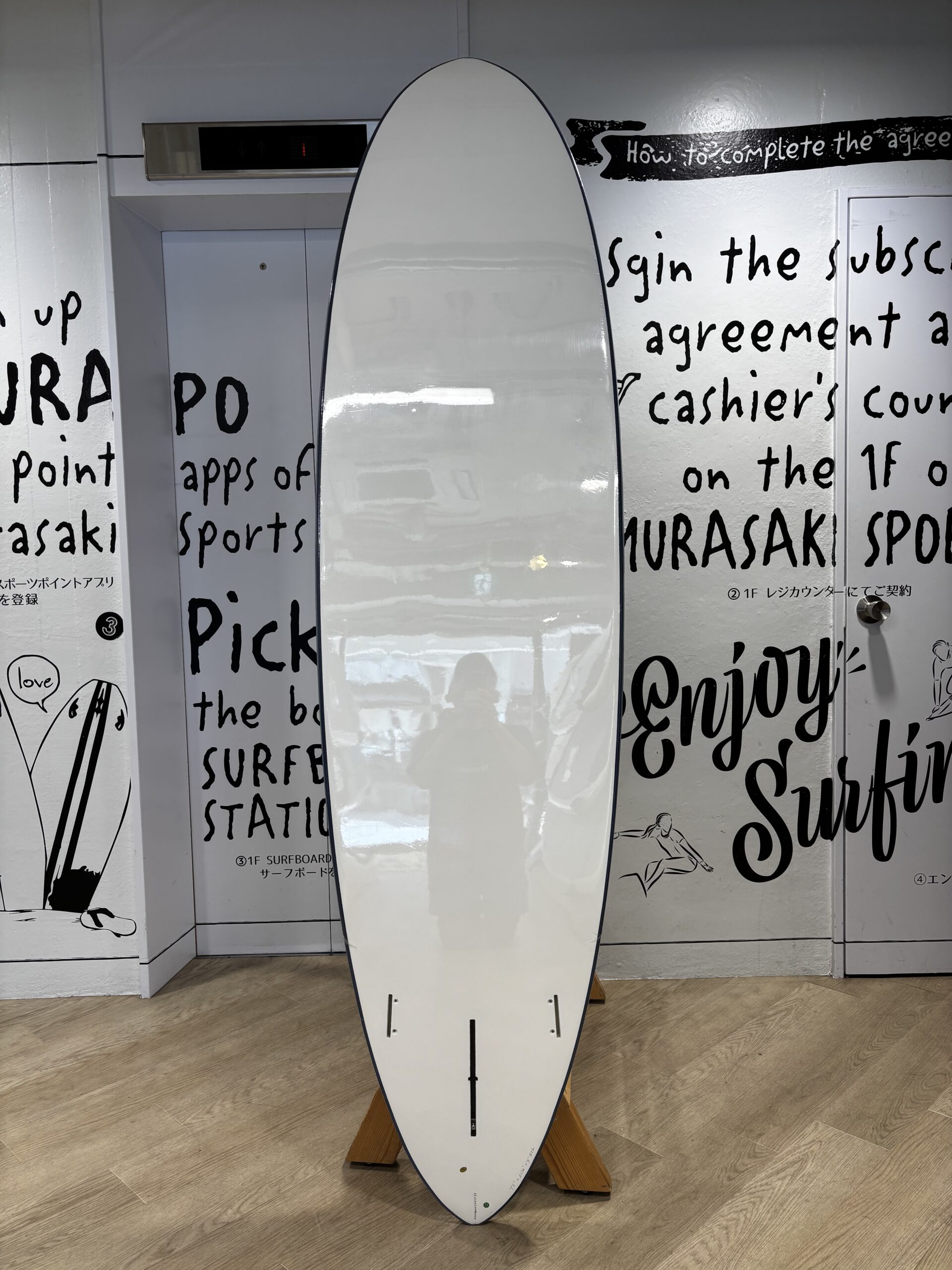 IMG_1133 - SHOP NEWS | ムラサキスポーツオンラインストア 通販 White surfboard on a wooden stand indoors, with a wall of black handwritten text and simple drawings behind it.