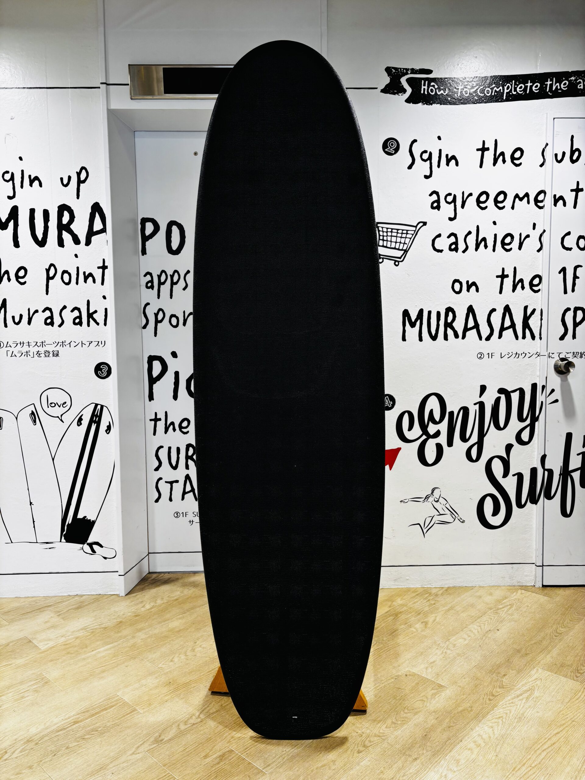 IMG_1143 - SHOP NEWS | ムラサキスポーツオンラインストア 通販 Vertical black surfboard-shaped object standing upright in a room with white walls covered in handwritten text and drawings, on a wooden floor.