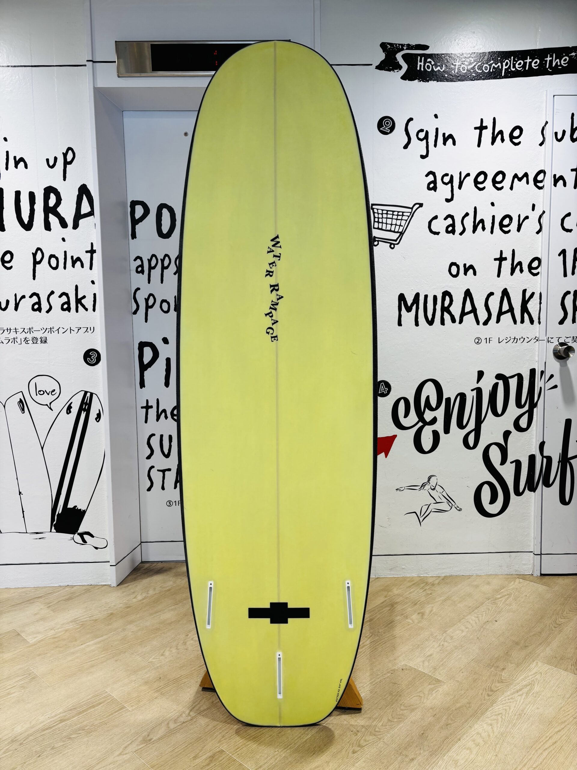 IMG_1144 - SHOP NEWS | ムラサキスポーツオンラインストア 通販 Yellow surfboard displayed indoors on a wooden floor, with bold black text graphics on white walls behind the board.