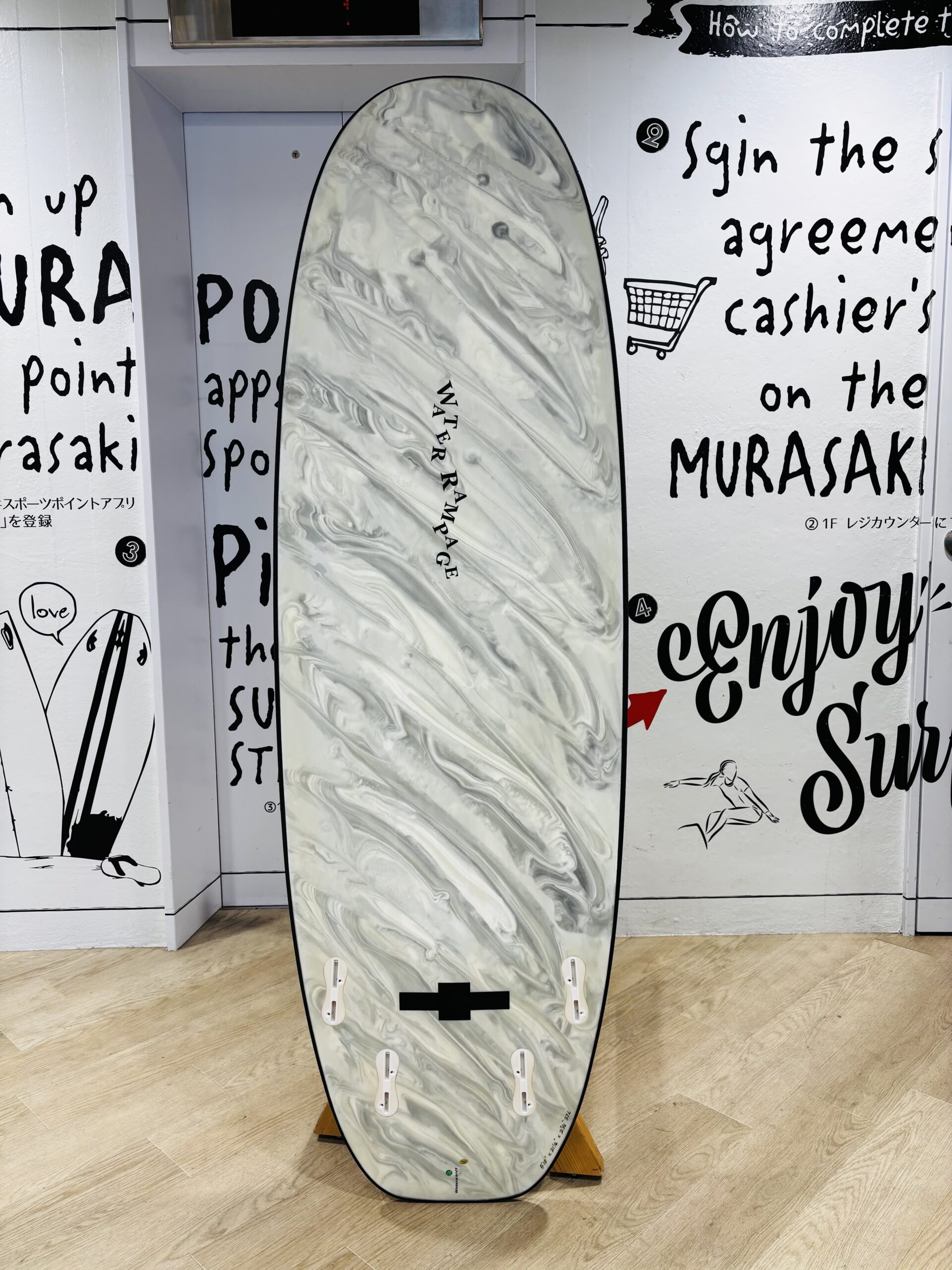 IMG_1146 - SHOP NEWS | ムラサキスポーツオンラインストア 通販 Marble-gray surfboard upright on a wooden stand inside a white-walled room with large black handwritten text on the walls; board has multiple fin boxes and 'WATER RAMPAGE' written along its center.
