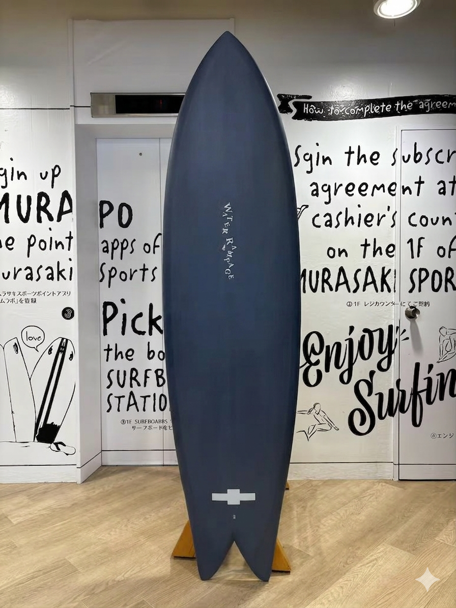 IMG_1147 - SHOP NEWS | ムラサキスポーツオンラインストア 通販 Blue surfboard on a wooden stand displayed upright in a shop, with bold wall art and text behind it.