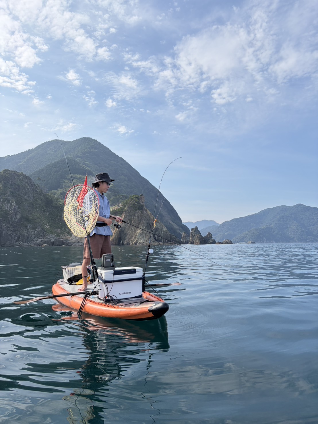 IMG_6738 - SHOP NEWS | ムラサキスポーツオンラインストア 通販 Person fishing on an orange inflatable paddleboard, standing with a rod against a backdrop of mountains and calm blue water.”