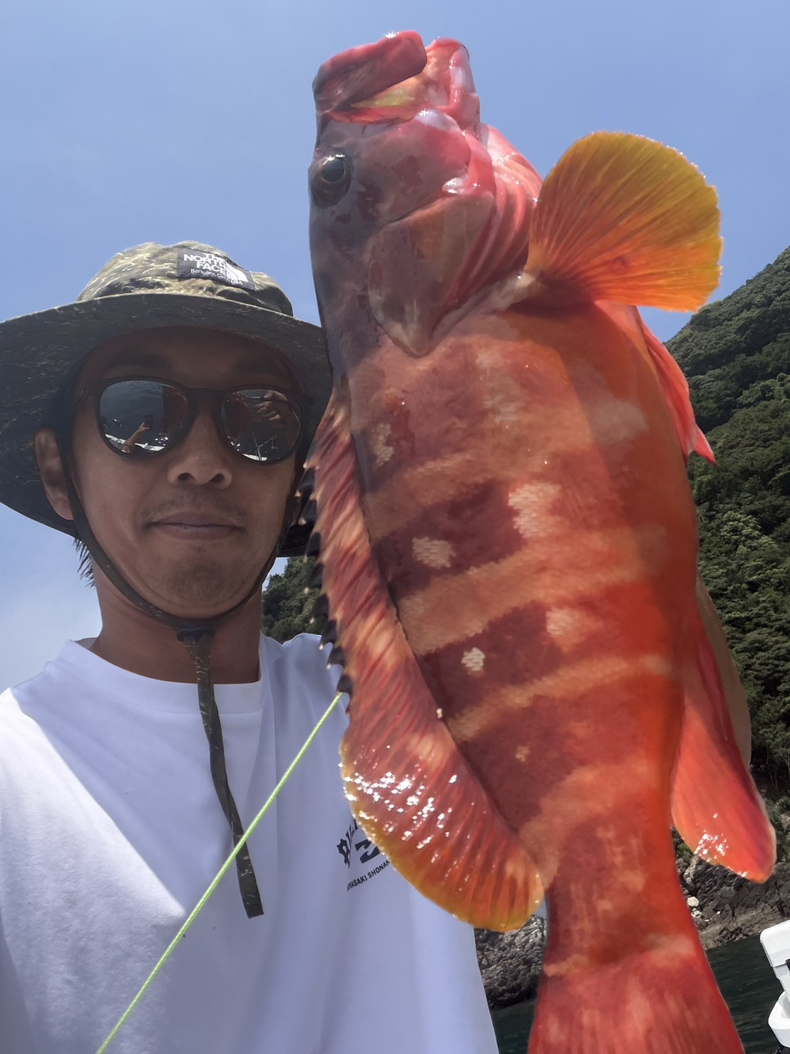 IMG_6739 - SHOP NEWS | ムラサキスポーツオンラインストア 通販 Man wearing a wide-brimmed hat and sunglasses holds a large red-orange fish up to the camera on a sunny day home shore.