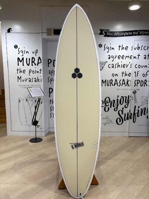 Cream-colored longboard surfboard on a display stand indoors, featuring a dark hexagon logo and a vertical stringer, set against graffiti-style walls in a showroom.