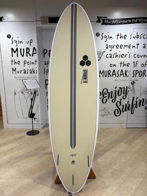 Cream surfboard with two dark grey stripes, upright on a stand in a shop, featuring a small hexagonal logo.