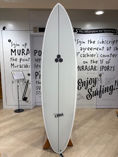 White surfboard on a wooden stand indoors, centered, with a small hexagonal logo along its stringer; graffiti-style wall and signage in the background.