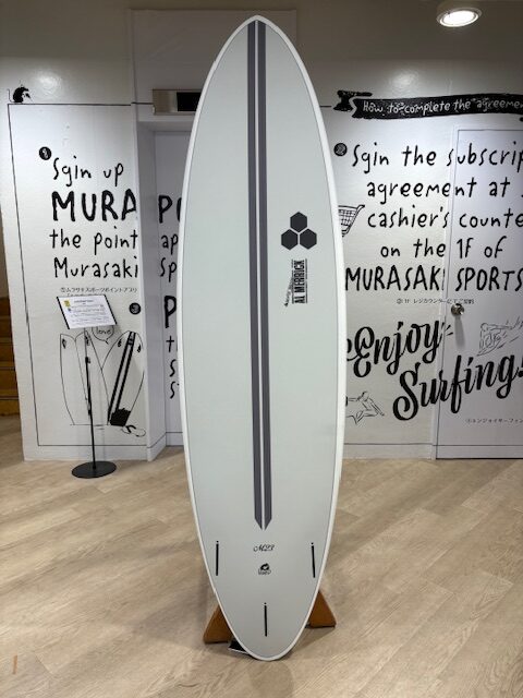 White surfboard on a wooden stand in a shop, featuring a dark gray center stripe and logo, with 'Enjoy Surfing' wall art in the background.