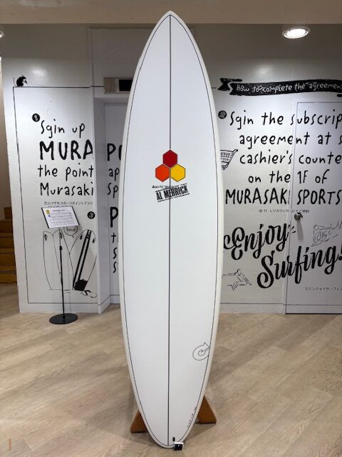 White surfboard with a red and yellow hexagonal logo centered, displayed upright on a wooden stand inside a surf shop with black graffiti-style wall art.