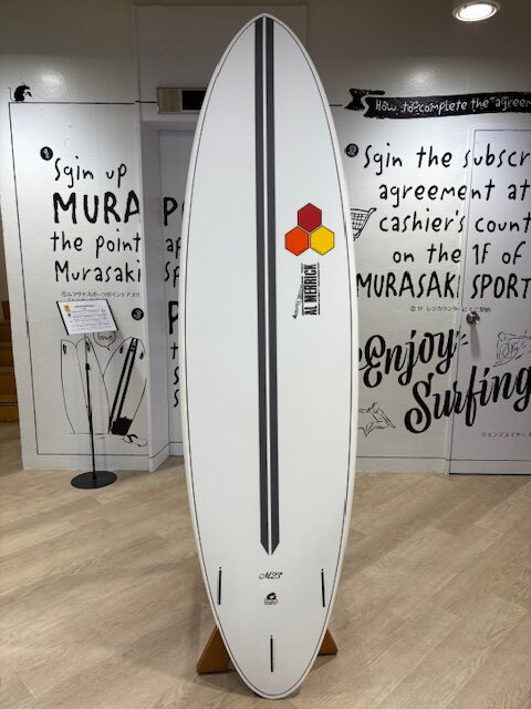 White surfboard with two dark gray center stripes stands on a wooden rack in a store; a hexagon logo and vertical label on the right side, with wall graphics and display stands in the background.