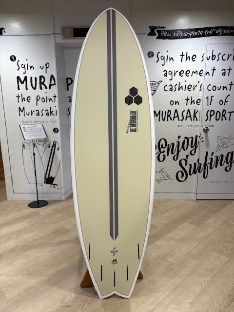 Cream surfboard with two dark gray stripes and a hexagonal logo, standing upright on a wooden rack in a surf shop showroom.