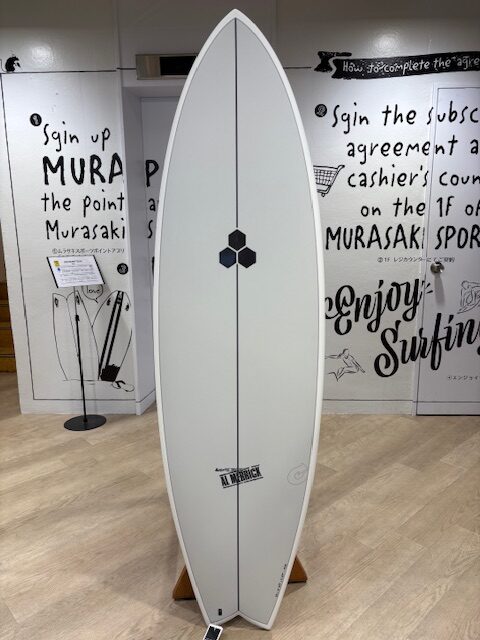 White surfboard on a stand indoors, featuring a vertical stringer, a small black hexagonal logo midboard, and a brand sticker near the tail.