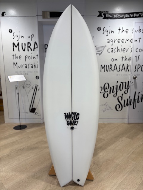 IMG_8801 - SHOP NEWS | ムラサキスポーツオンラインストア 通販 White surfboard on a wooden stand in a showroom, with a 'Magic Surf' logo toward the bottom and graffiti-style walls behind.