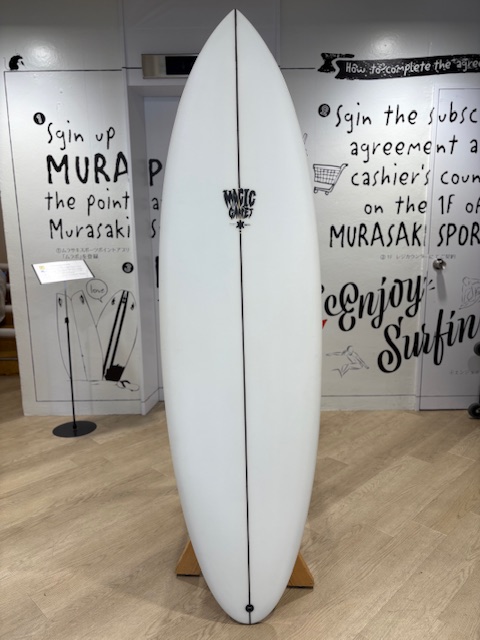 IMG_8808 - SHOP NEWS | ムラサキスポーツオンラインストア 通販 White surfboard on a wooden stand indoors, featuring a dark stringer and a small black logo near the center.