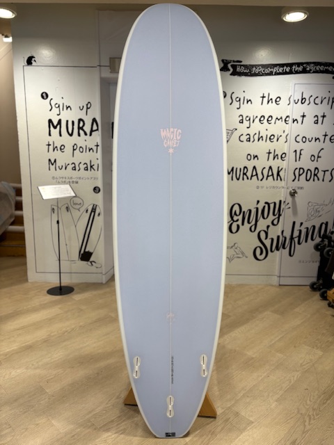 IMG_8831 - SHOP NEWS | ムラサキスポーツオンラインストア 通販 Light blue longboard on a wooden display stand in a surf shop, with handwritten wall art about surfing in the background.