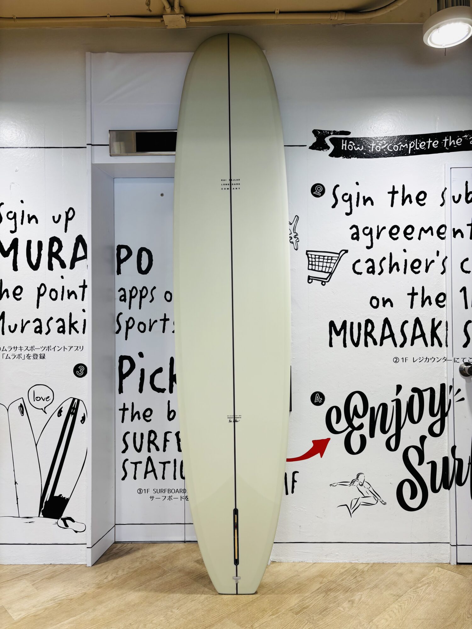 Cream-colored surfboard standing upright against a white wall with bold black graffiti text indoors, with warm overhead lighting and a wooden floor.