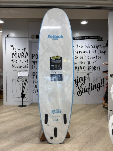IMG_9654 - SHOP NEWS | ムラサキスポーツオンラインストア 通販 White surfboard with blue edge on a display rack in a shop, Softech logo near the top and a product label in the center.