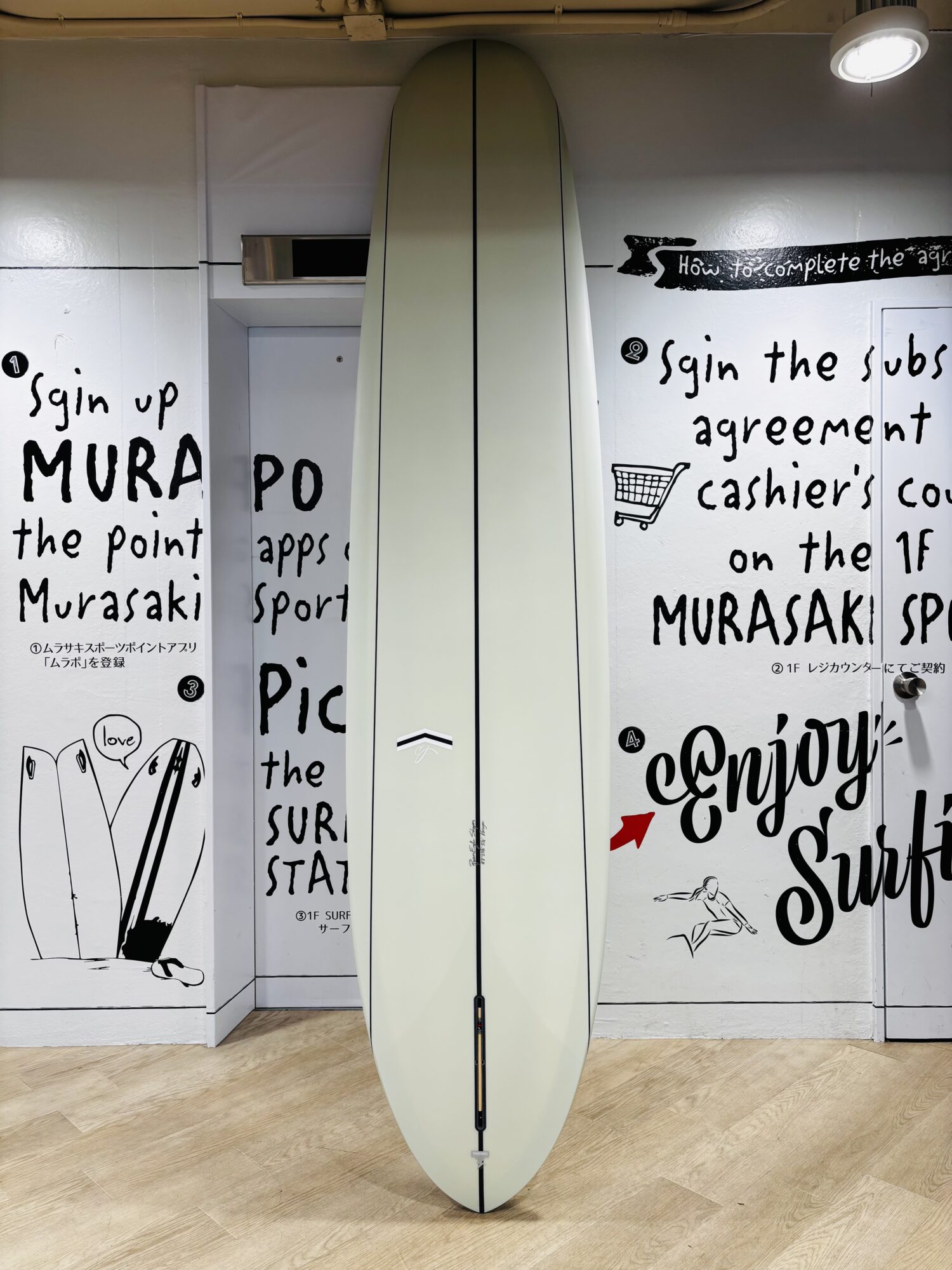 Cream surfboard standing upright in a room with white walls covered in black handwritten graffiti and drawings. The wooden floor is visible at the bottom.