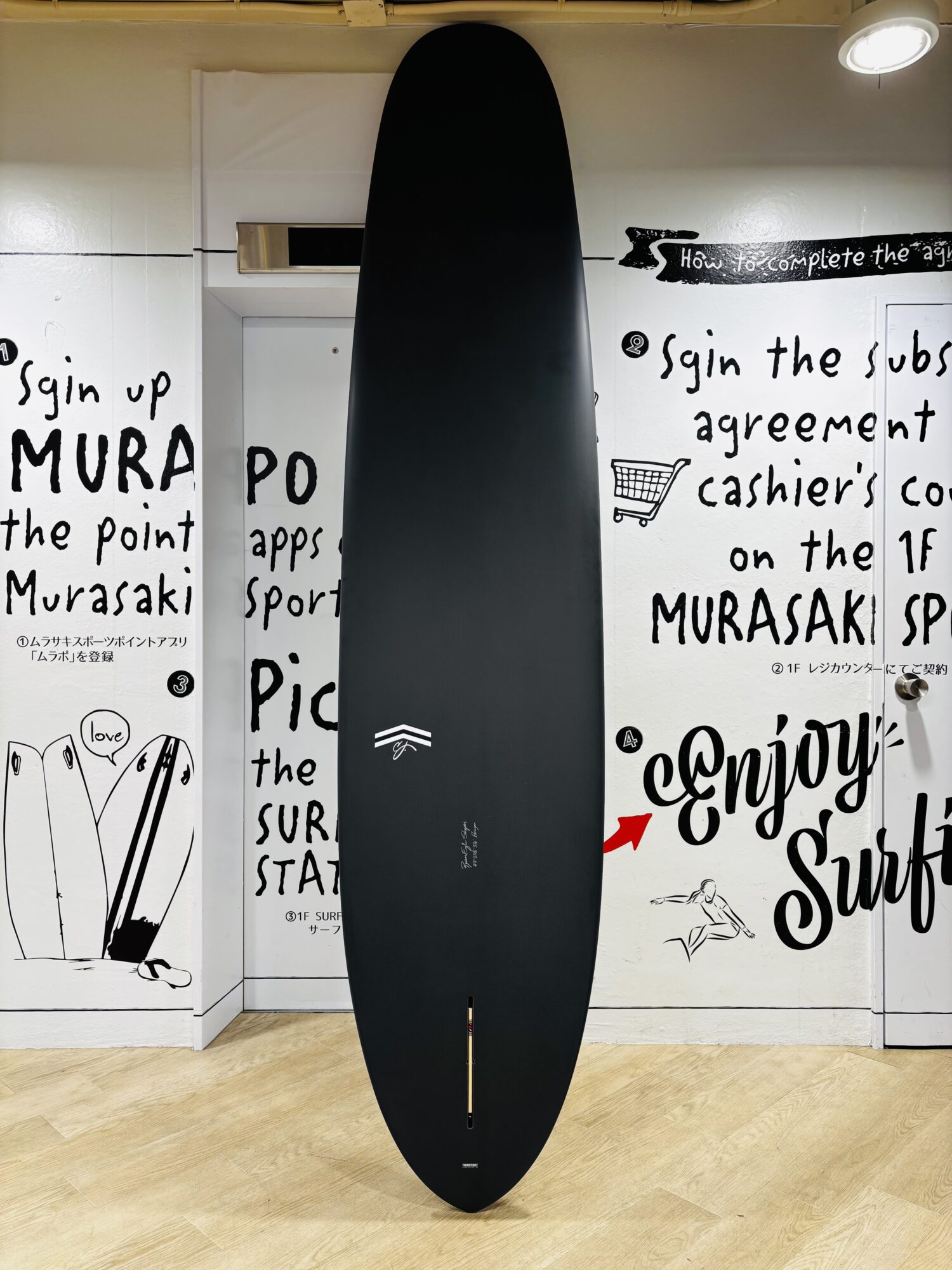 Vertical black surfboard stood upright in a showroom with white graffiti-covered walls and wooden floor.