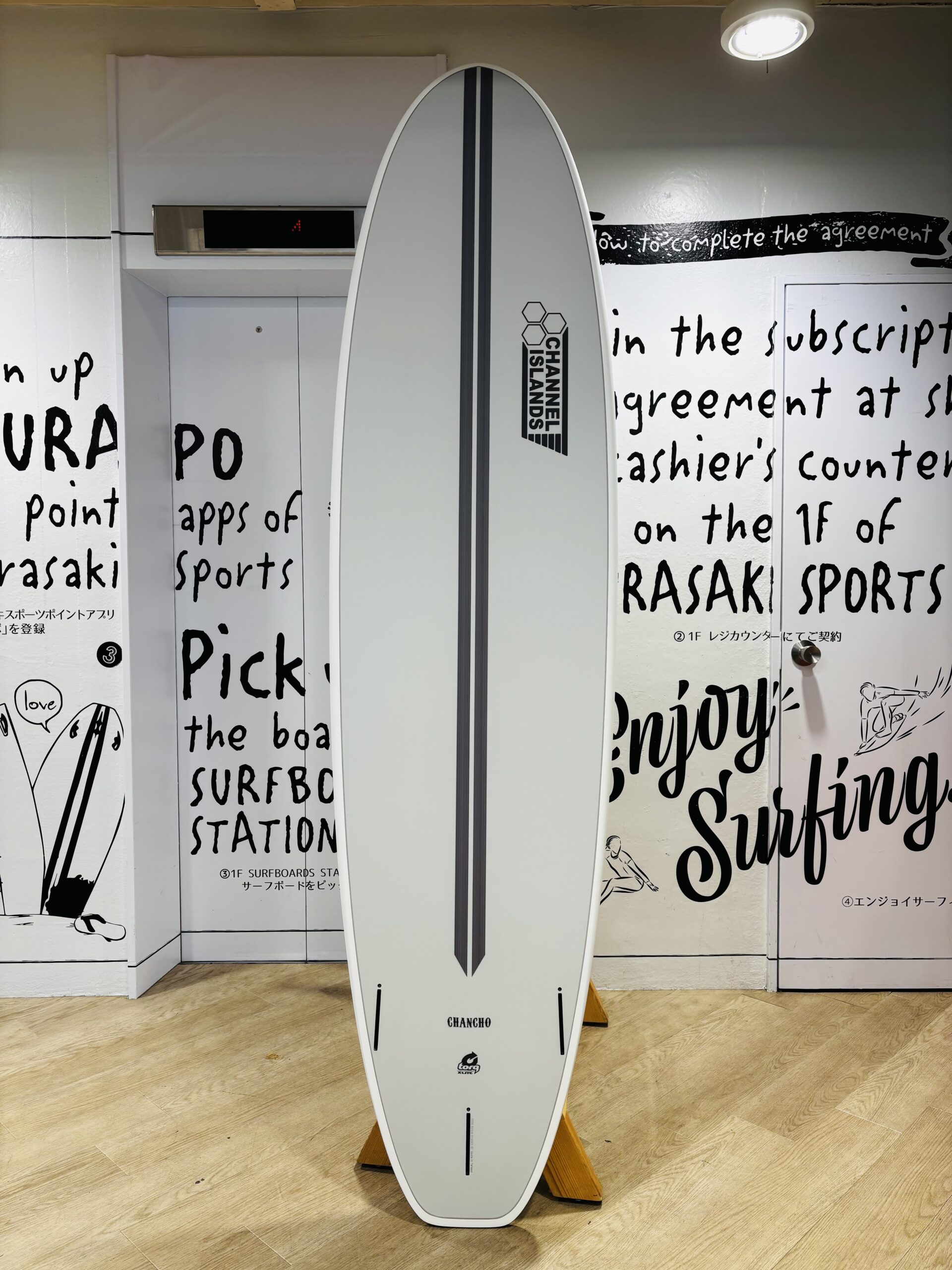 IMG_9969 - SHOP NEWS | ムラサキスポーツオンラインストア 通販 Tall white surfboard on a yellow stand indoors, with a Channel Islands logo, set against walls covered in large black script text.