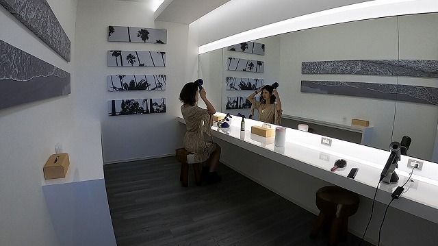 Woman at a bright white makeup vanity applying makeup while looking in the large mirror; wall shelves display black-and-white palm tree photos.