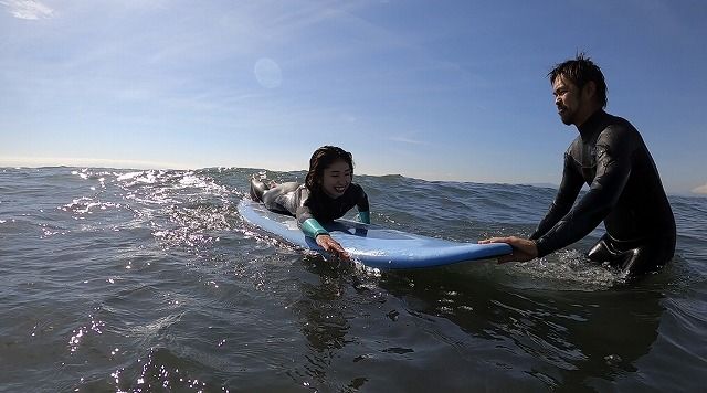 A young girl lies on a blue surfboard riding a small wave while a man in a wetsuit supports the board in the water next to her.