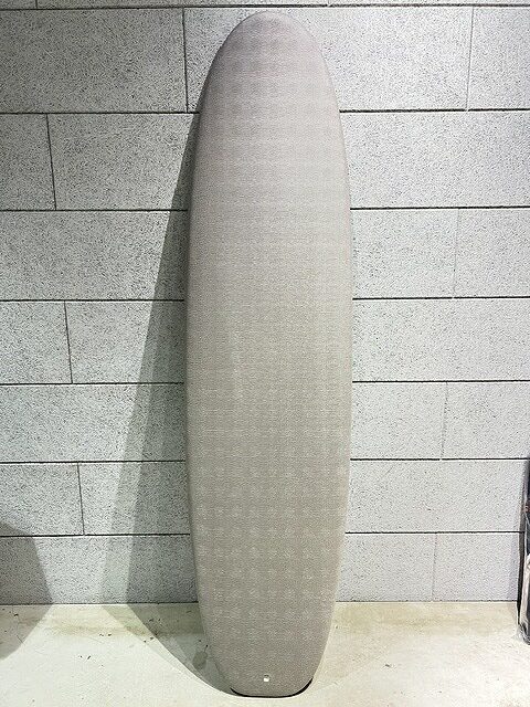s-IMG_7411 - SHOP NEWS | ムラサキスポーツオンラインストア 通販 Light gray surfboard with rounded nose standing upright against a gray brick wall area.