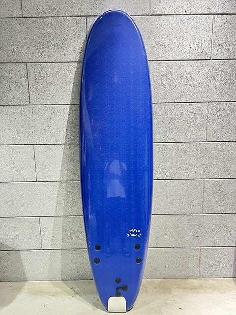s-IMG_7417 - SHOP NEWS | ムラサキスポーツオンラインストア 通販 Blue surfboard standing upright against a light gray brick wall inside an indoor space.