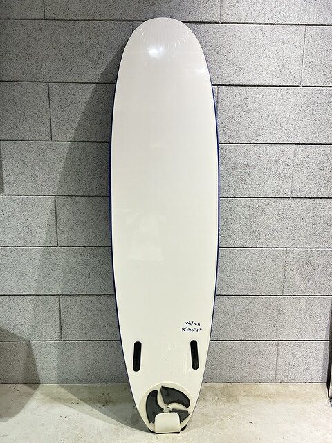 s-IMG_7419 - SHOP NEWS | ムラサキスポーツオンラインストア 通販 White surfboard with a rounded nose standing upright against a gray brick wall; two small fin slots near the tail and a single fin box visible at the bottom area