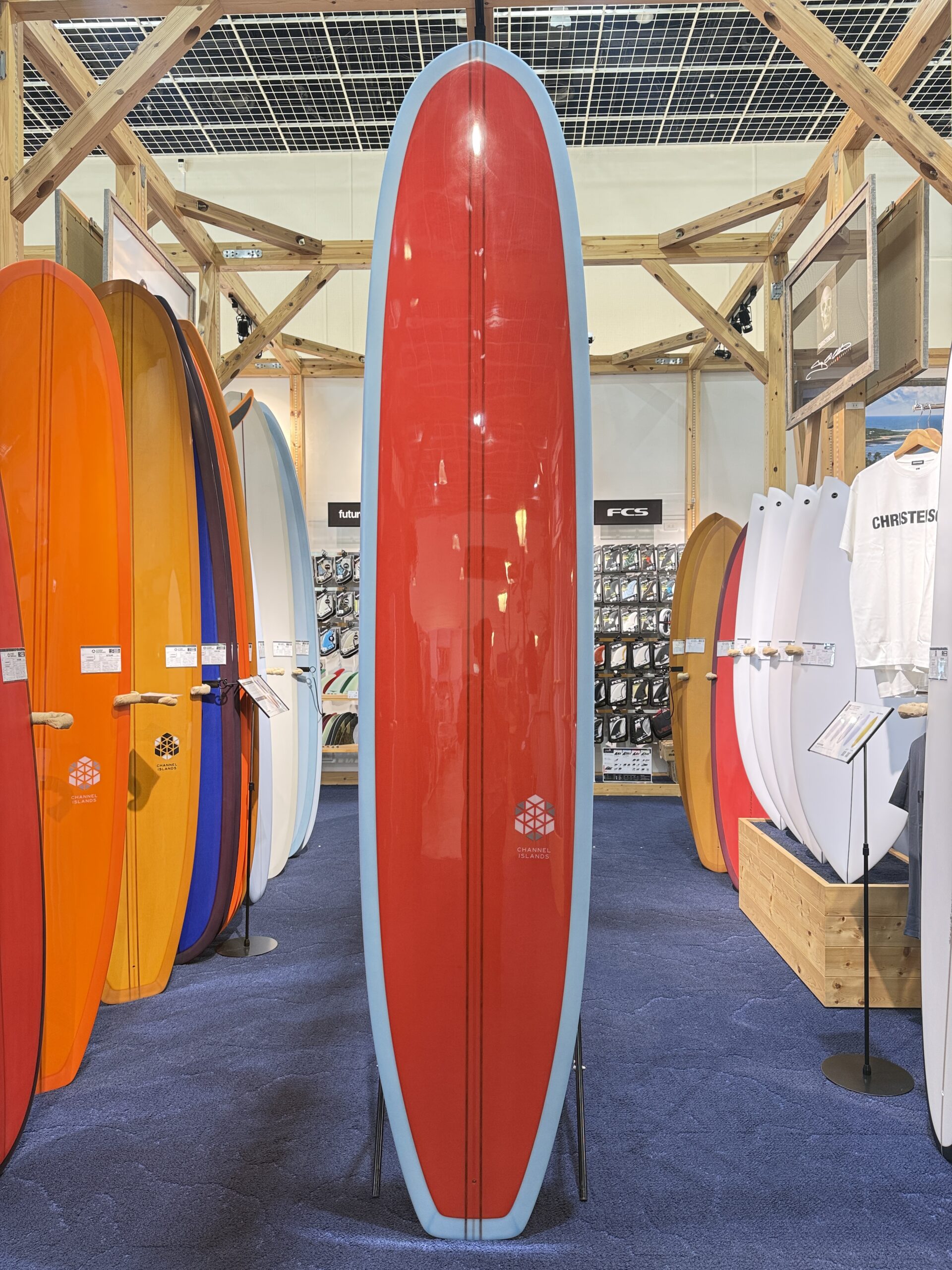 Tall red surfboard with light blue edge standing upright in a store display among orange, yellow, and white boards.