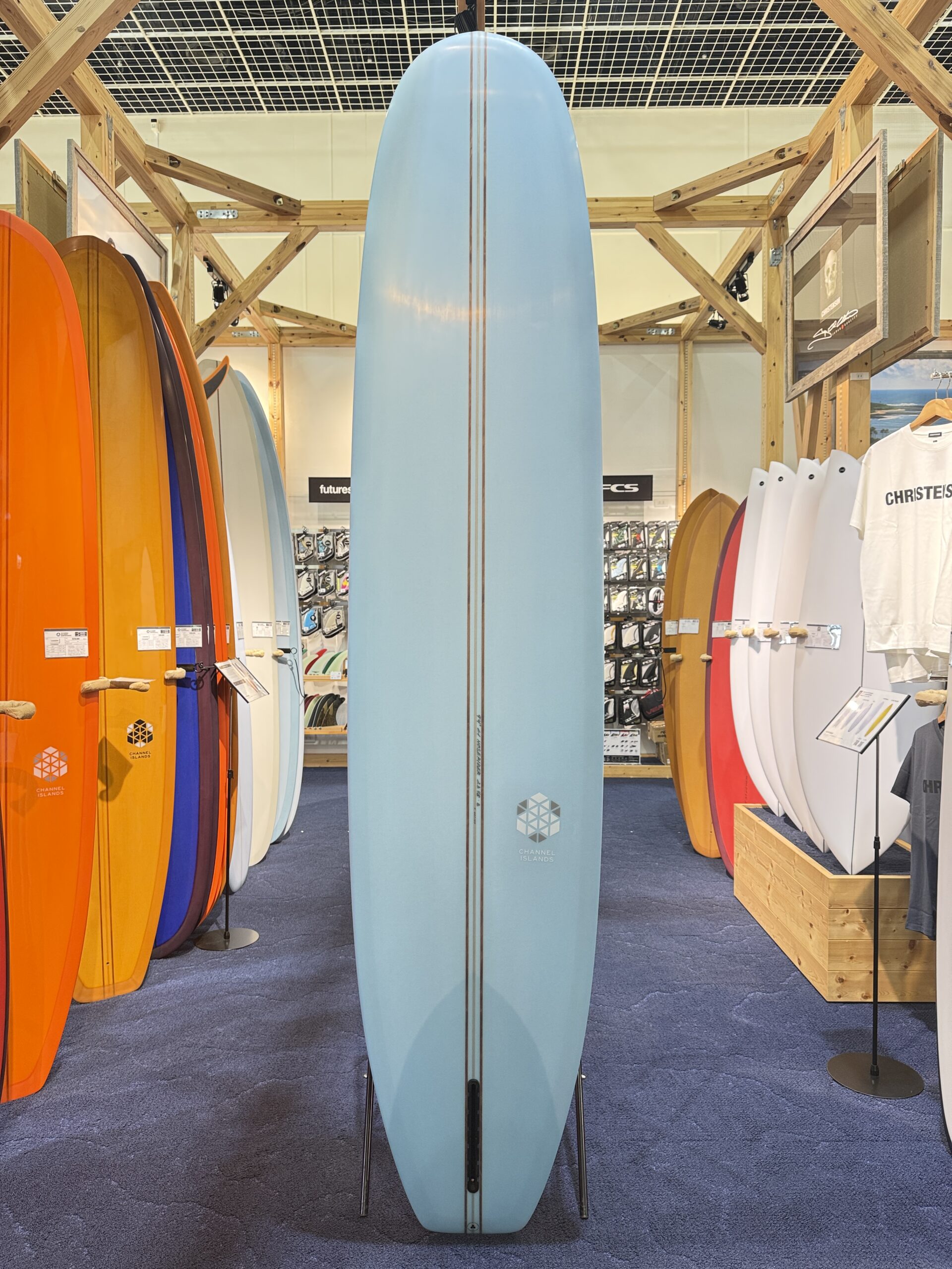 Center blue longboard with brown pinstripes stands upright on metal legs in a surf shop, surrounded by colorful boards and wooden display racks, with the Channel Islands logo toward the base.