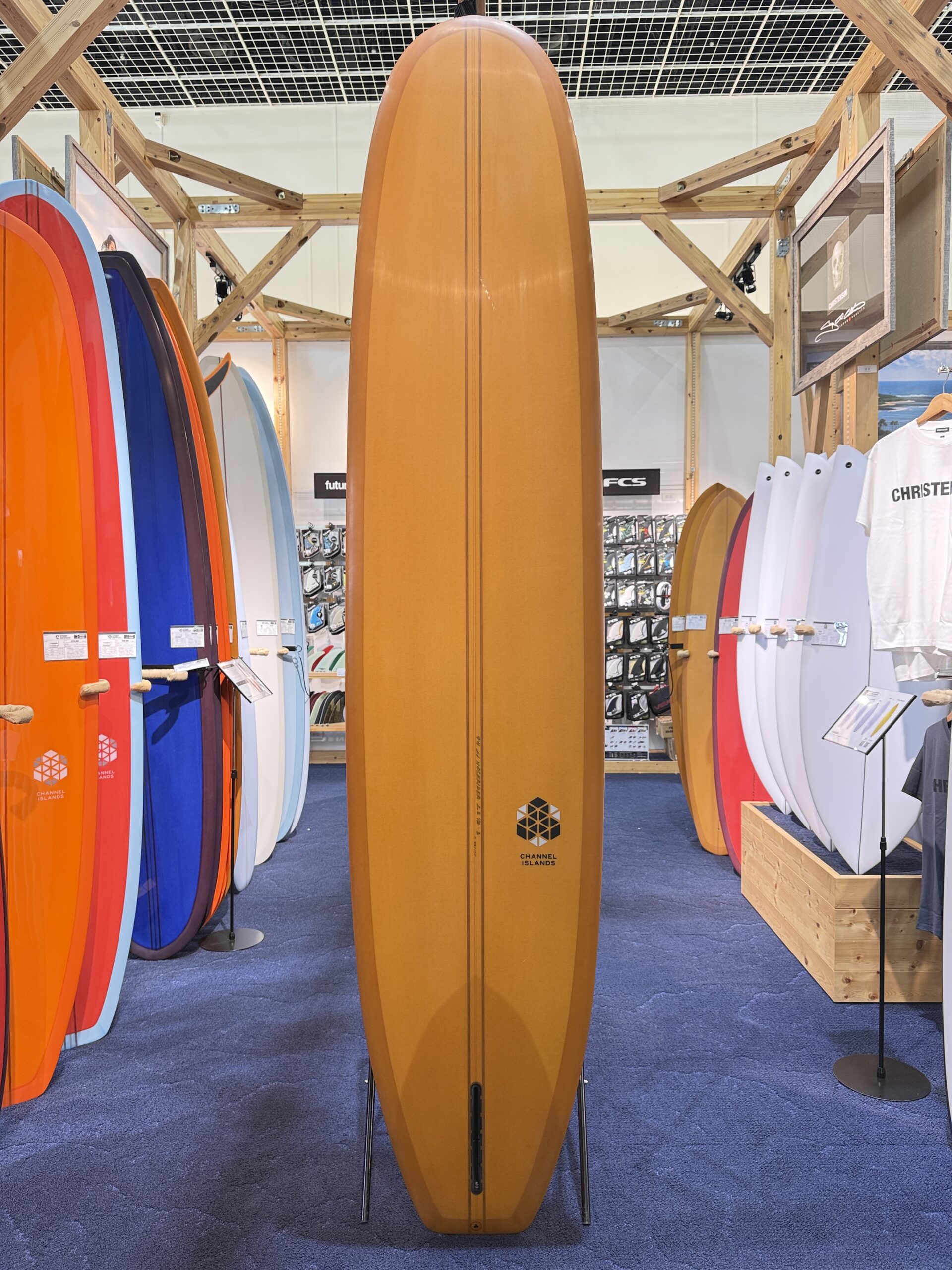 Tall orange surfboard standing upright in a surf shop display, surrounded by colorful boards on racks and a wooden frame structure behind it.