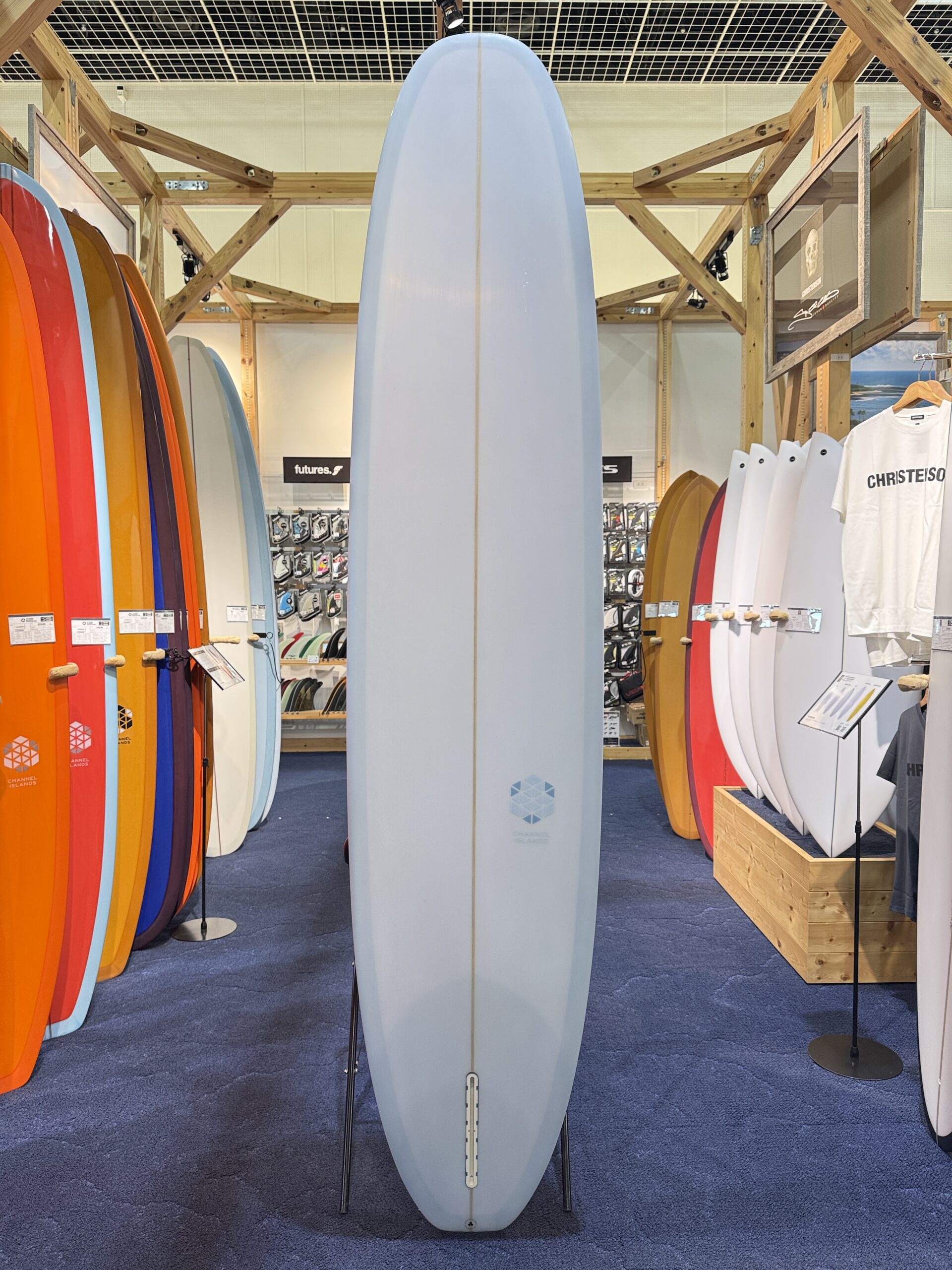 Tall light-blue surfboard on a stand in a crowded surf shop, with colorful boards lining the sides and white ones on the right.