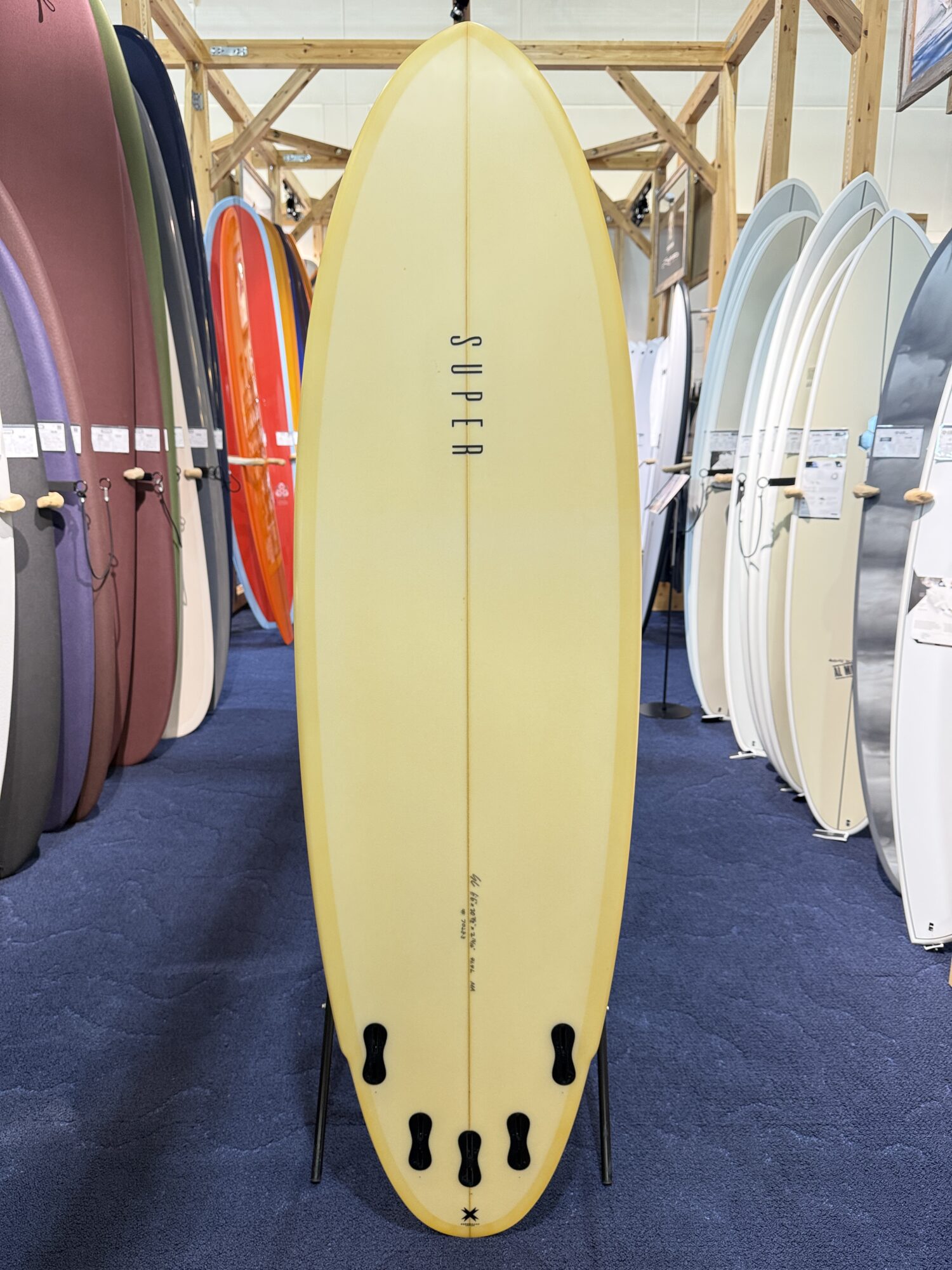 Yellow surfboard with four fins on a stand in a surf shop, flanked by colorful boards on racks.
