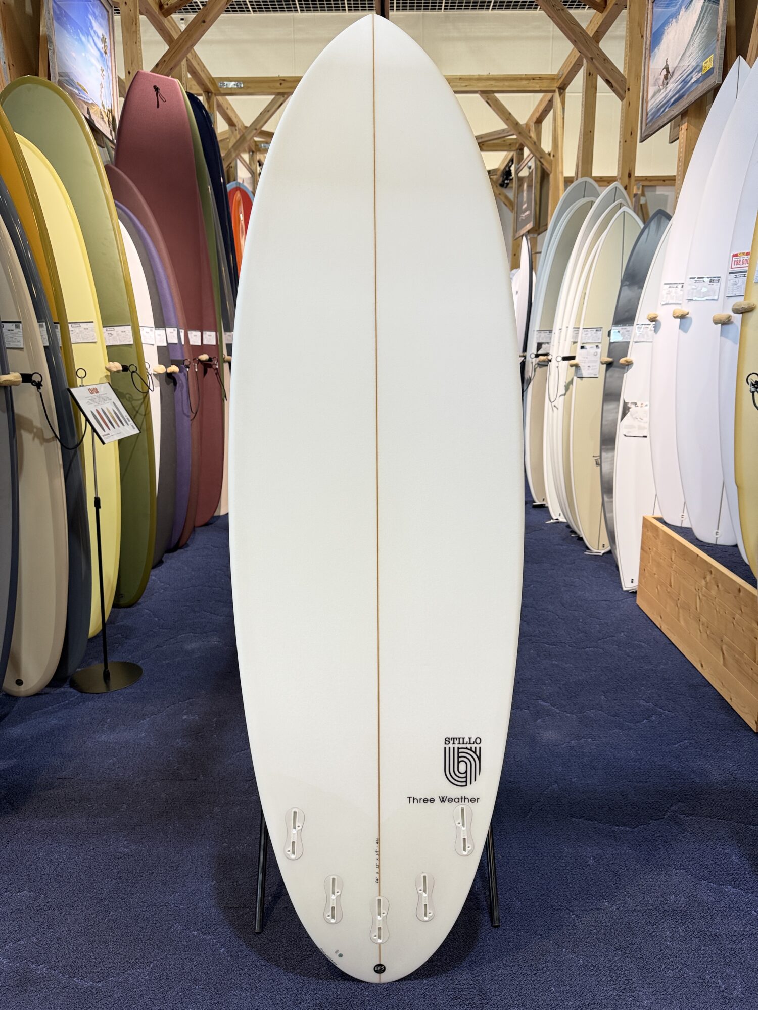 IMG_4020 - SHOP NEWS | ムラサキスポーツオンラインストア 通販 White surfboard standing upright in a store, surrounded by colorful boards on racks; wooden beams overhead, blue carpeted floor, logo and 'Three Weather' text near the tail.