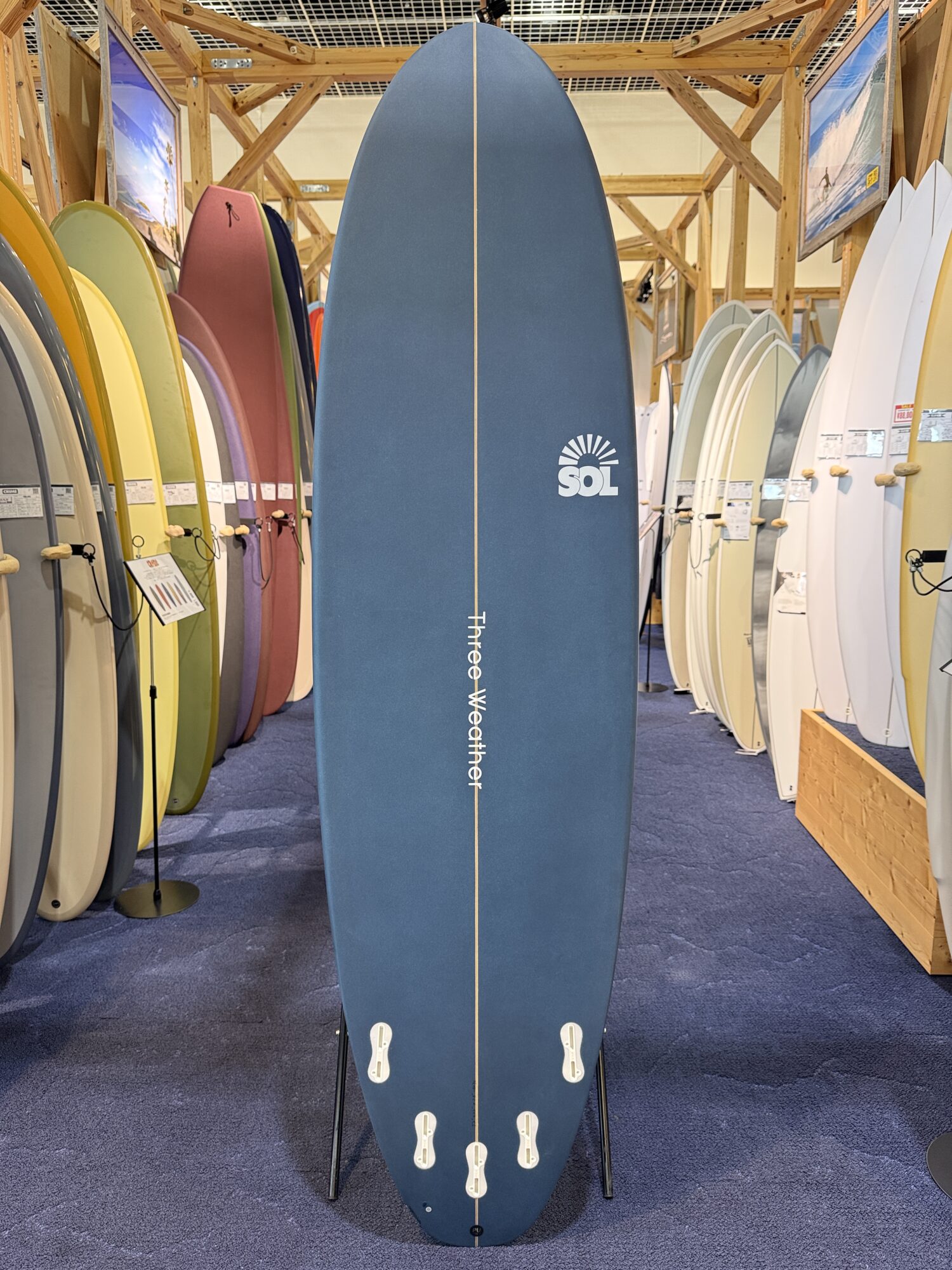 IMG_4028 - SHOP NEWS | ムラサキスポーツオンラインストア 通販 Large blue surfboard on a stand in a store, with white fins at the bottom and a vertical stringer running down the center, surrounded by colorful boards on display.