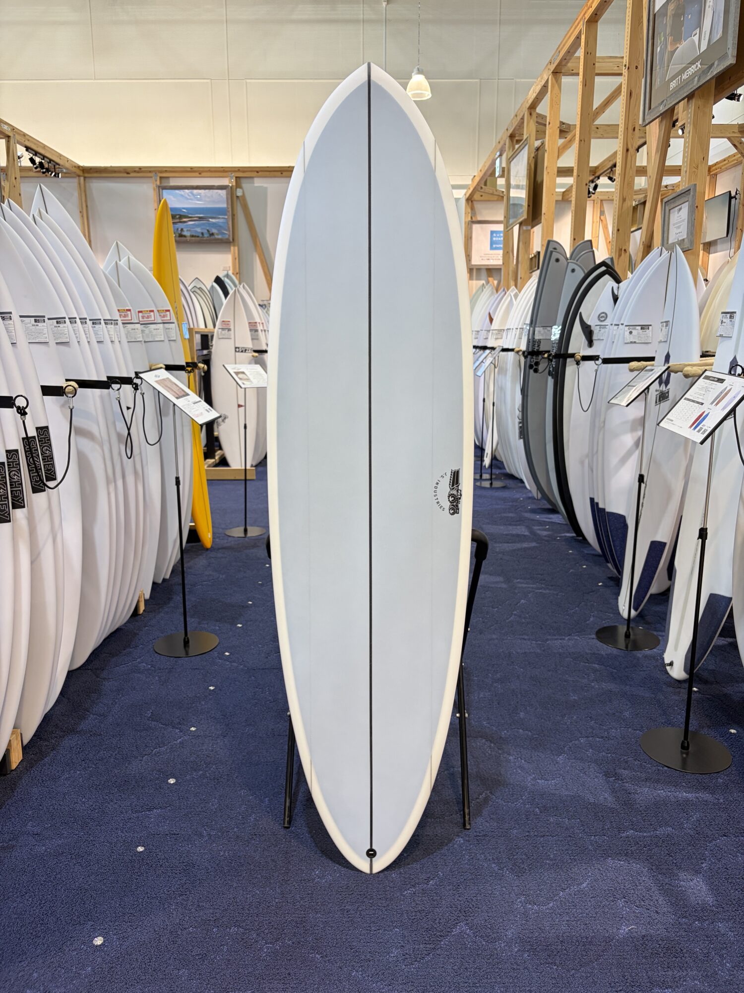 Central light-blue surfboard on display stand in a showroom, surrounded by rows of white surfboards.
