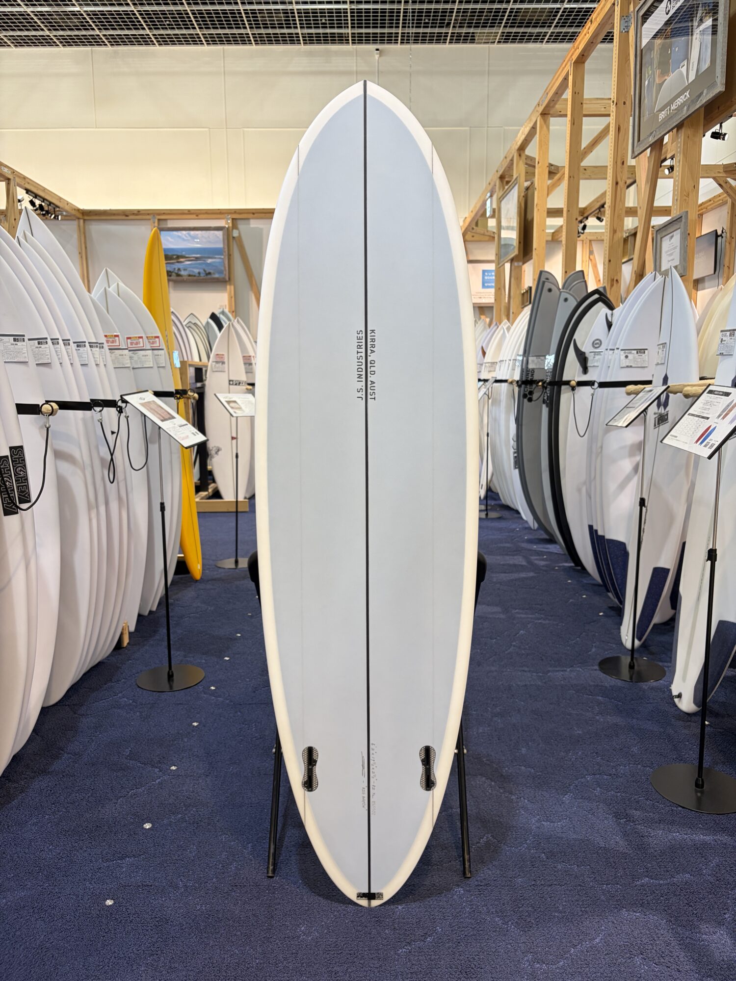 Tall, pale blue surfboard on a stand in a showroom with rows of white and gray boards around it.