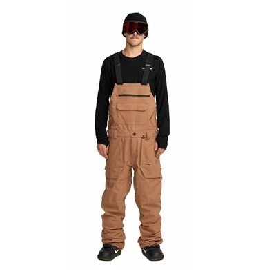 Man in tan snowboard bib overalls with black long-sleeve shirt and red goggles.
