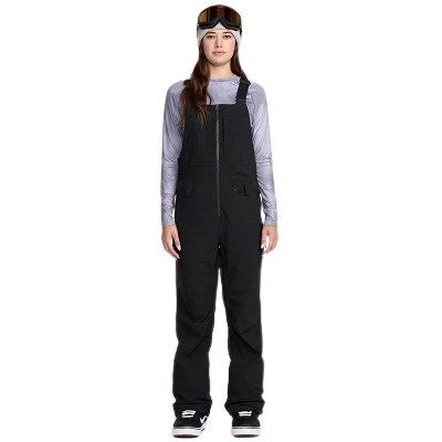 Full-body view of a person in black ski bib overalls with a gray long-sleeve shirt, goggles on their head, and sneakers, facing forward.