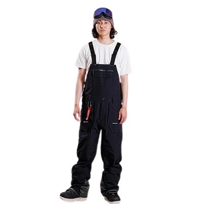 Young person standing in black bib overalls over a white T-shirt, with blue goggles on their head, on a white background.