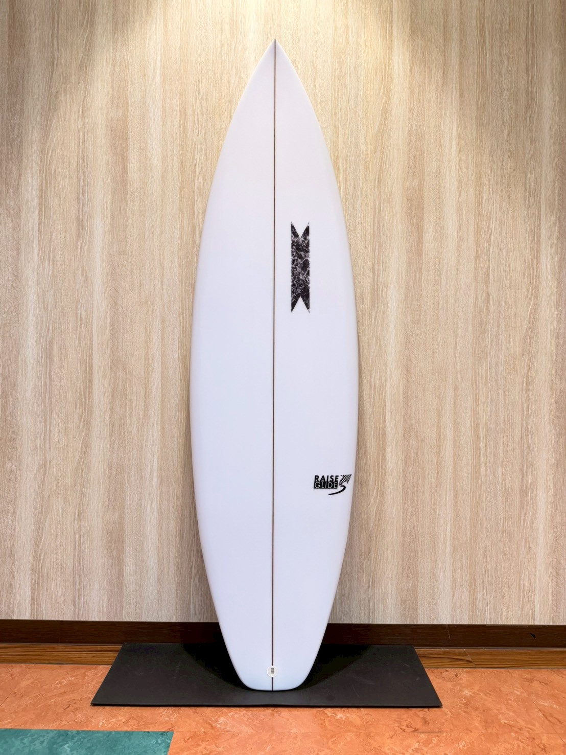 LINE_ALBUM_2026 SB_260427_1 - SHOP NEWS | ムラサキスポーツオンラインストア 通販 White surfboard standing upright on a black mat against a wood-paneled wall, with a small black logo near the bottom.