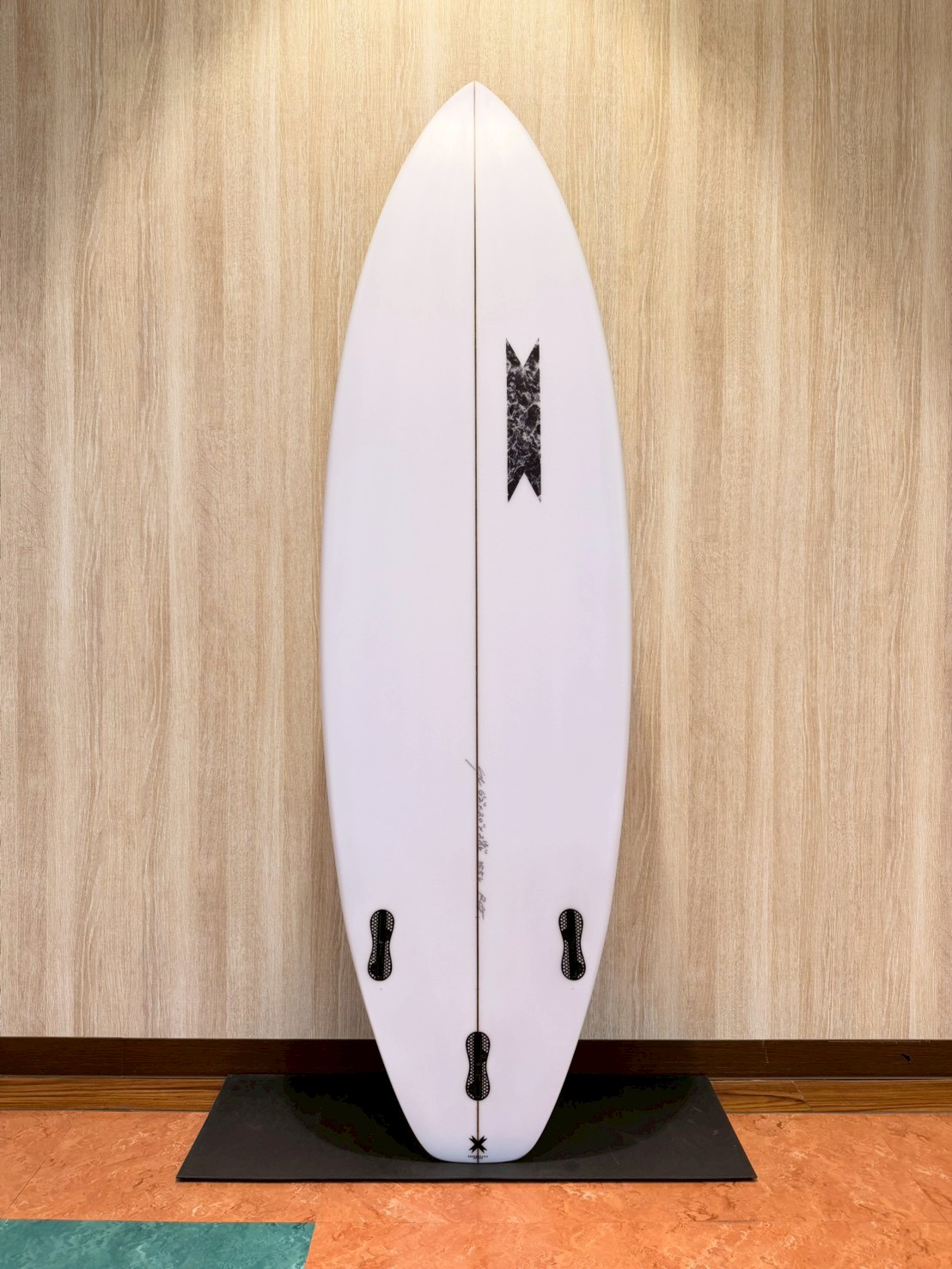LINE_ALBUM_2026 SB_260427_2 - SHOP NEWS | ムラサキスポーツオンラインストア 通販 White surfboard standing upright on a black display stand against a wood-paneled wall; fin boxes visible along the rails, minimal branding near center.