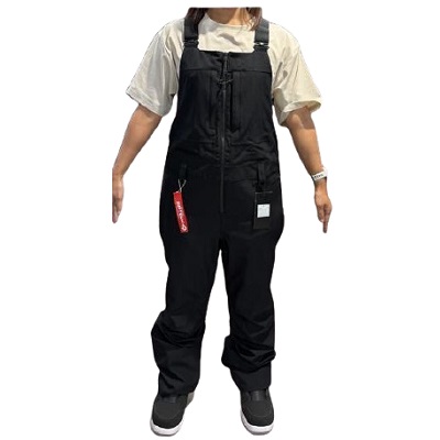 Person wearing black bib overalls with a light beige T-shirt, standing with arms by sides; red tag hanging from the zipper pocket.