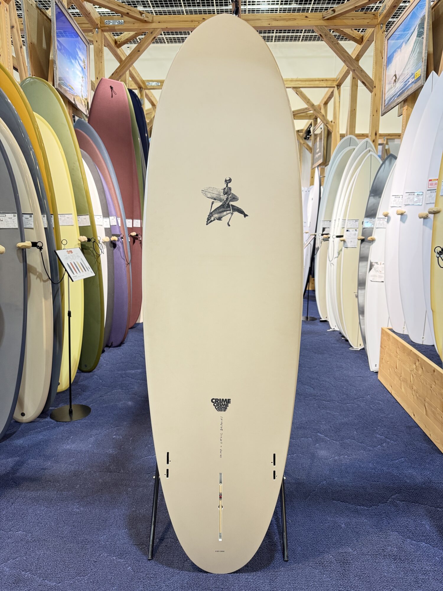 IMG_4050 - SHOP NEWS | ムラサキスポーツオンラインストア 通販 Beige surfboard on a stand in a shop aisle, surrounded by colorful boards on racks and wooden framing in the background.